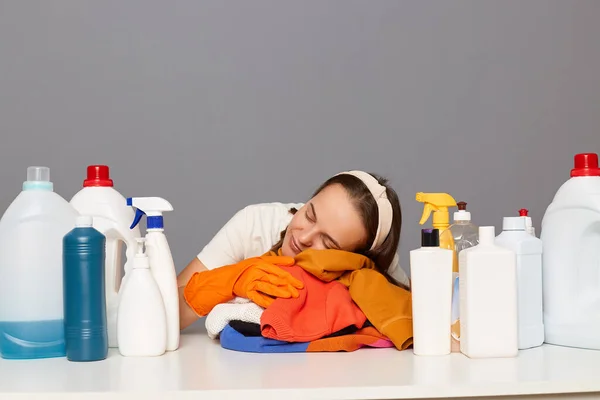 Tired sleepy Caucasian woman wears rubber protective gloves and headband, surrounded with bottles of detergents for laundry, leaning on table, sleeping, feels tired and exhausted.
