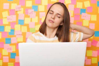 Portrait of tired sad sleepy woman wearing striped T-shirt posing over sticky notes to write reminder on yellow wall, working on pc computer, being overworked, feels pain in neck.