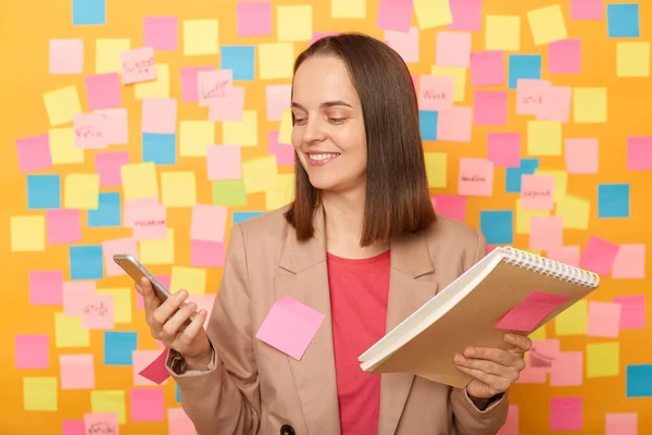 Portrait of smiling joyful delighted brown haired woman wearing beige jacket, using cell phone for checking e-mail, working with papers, posing against colorful sticky notes on yellow wall.