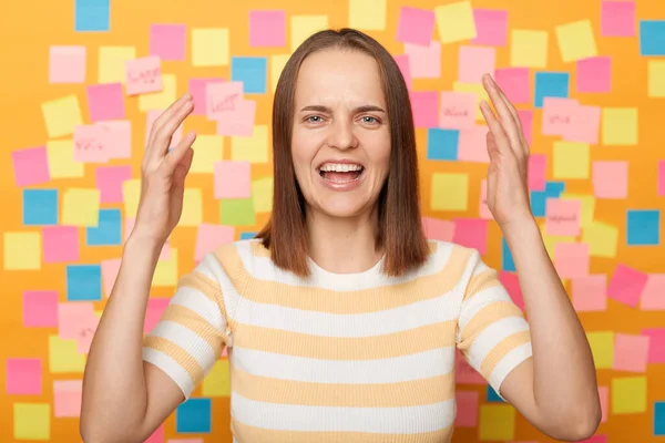 Photo of shocked sad despair brown haired woman wearing T-shirt, having serious problems, raised arms and screaming, posing against colorful sticky notes on yellow wall.