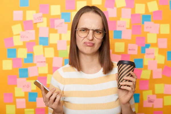 Photo of sad upset depressed brown haired woman wearing T-shirt posing against colorful sticky notes on yellow wall, drinking coffee, using mobile phone, frowning face and looking at camera.