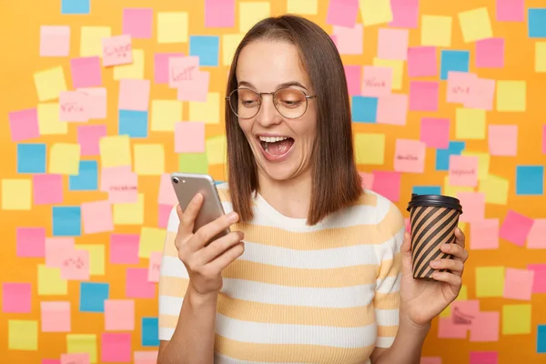 Portrait of excited amazed happy woman wearing striped T-shirt posing over sticky notes to write reminder on yellow wall, drinking coffee, using mobile phone, reading good news.