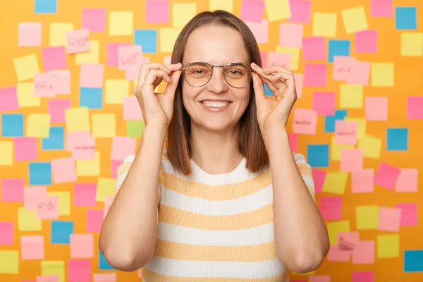 Horizontal shot of smiling satisfied attractive young adult woman wearing T-shirt posing against yellow paper wall with adhesive notes, keeps hand on frame of her glasses and looking at camera.