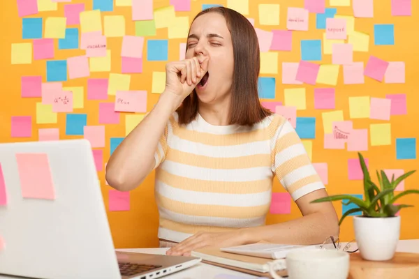 Photo of sleepy brown haired woman wearing T-shirt posing against colorful sticky notes on yellow wall, working on laptop, feels sleepy, yawning while sitting in front of computer.