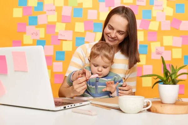 Indoor shot of charming dark haired woman wearing striped t-shirt sitting at table, posing against memo cards on yellow wall, female combines maternity leave with her online job, playing with kid.