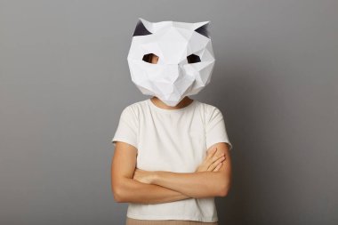 Kapalı alanda, kedi maskesi ve beyaz tişört giyen gururlu bir kadının gri arka planda izole edilmiş poz verdiği, elleri bağlı duran, kendine güvenini ifade eden bir fotoğraf..