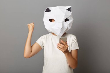 Neşeli, mutlu, kedi maskesi takan, beyaz tişört giyen, cep telefonu kullanan, yumruğunu sıkan, zaferi kutlayan, gri arka planda izole edilmiş poz veren mutlu kadının fotoğrafı..