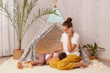 Güzel kahverengi saçlı bir kadının, çocukları çadırın yanında otururken ve cep telefonuyla konuşurken kızı bacaklarının üzerinde uyurken, bir kadının sohbet ederken görüntüsü..