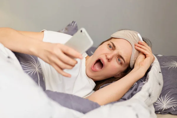 Photo of woman in bed with sleep mask wakes up and looking at mobile display, sees unexpected news on a smartphone early in the morning, being oversleep, screaming, keeps mouth opened.