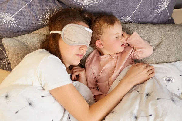 Indoor shot of woman in sleeping mask lying with her baby daughter and sleeping, family lying under blanket, female in sleeping mask, hugging her daughter.