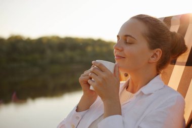 Nehir kıyısındaki güverte sandalyesinde dinlenen, el ele tutuşan, hoş bir kahve aromatı kokan, beyaz gömlekli sakin genç bir kadının yan görüş portresi..