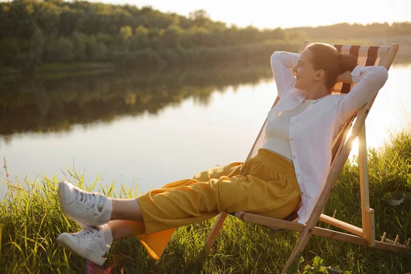 Full length of joyful cheerful woman with bun hairstyle relaxing on a deck chair on the river bank, raised her rams behind head, having relaxed look, enjoying beautiful nature and her recreation.