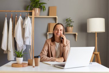 Ofiste bej ceket giyen, kameraya bakan, mutlu bir şekilde gülümseyen, iş ortağıyla telefonda konuşan güzel beyaz kadın tasarımcının fotoğrafı..