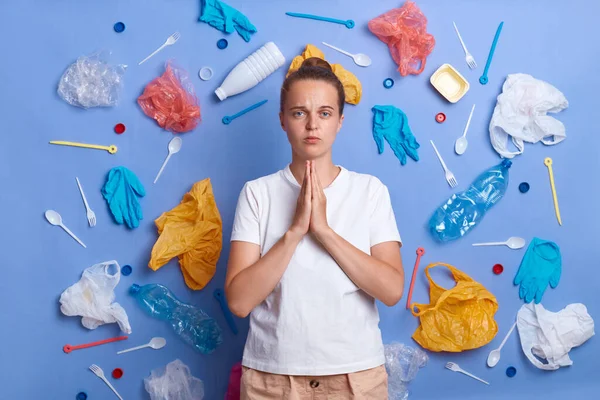 Portrait of sad unhappy woman dressed in white T-shirt, picked plastic litter isolated on blue wall, standing with palms together, praying for Earth environment, looks at camera with upset expression.