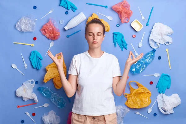 Portrait of calm woman dressed in white T-shirt, picked plastic litter isolated on blue wall, practicing yoga, trying to calm down, worries about the environment and causing harm to nature.