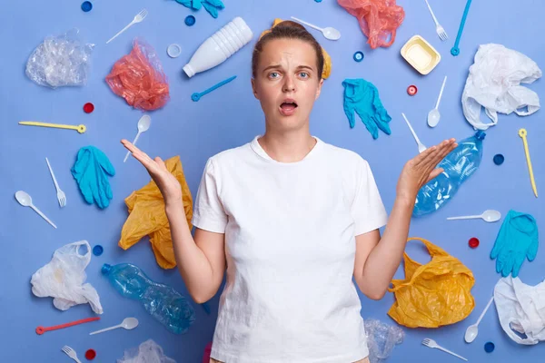 Image of shocked sad woman dressed in white T-shirt, picked plastic litter isolated on blue wall, raised arms, asking how can we stop it,wants to live on clean Earth, looking at camera.
