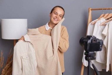 Cute charming satisfied smiling woman wearing beige jumper, showing warm soft sweater, creating content for her vlog, recording video, standing against gray wall with clothes on hangers on shelf.