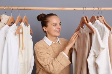 Image of attractive positive woman wearing beige sweater, posing in shopping mall, choosing new outfit, enjoying shopping, standing against gray wall with clothes hang in wardrobe on rack.