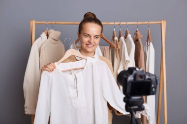 Cheerful smiling woman with bun hairstyle wearing beige jumper standing against gray wall with clothes on hangers on shelf, showing white stylish shirt to camera, shooting video for vogue vlog.