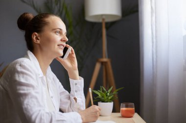 Side view portrait of dark haired adorable smiling woman with brown hair, sitting at table and talking on smart phone, looking away, holding pencil in hand, writing notes.