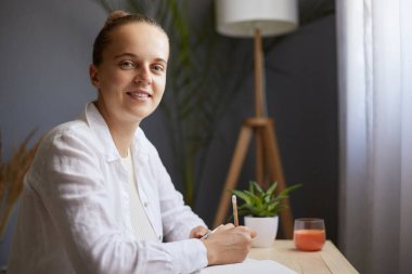 Indoor shot of attractive beautiful smiling young woman writing down notes while sitting at table in office, looking at camera with positive expression, wearing casual clothing.