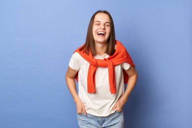 Indoor shot of joyful delighted Caucasian girl wearing white T-shirt and orange sweater tied over shoulders, looking at camera, laughing, hearing joke, standing against blue wall.