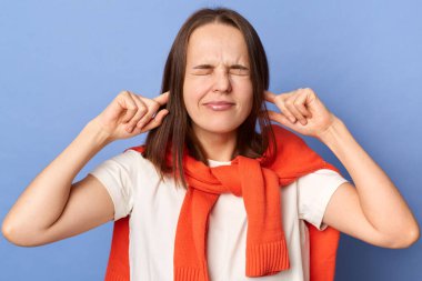 Frustrated annoyed woman in T-shirt and sweater tied over shoulders, covering ears with hands, feeling irritated with loud annoying noise, can't concentrate on her work, isolated on blue background.