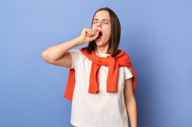 Indoor shot of sleepless tired exhausted woman dressed white T-shirt and tied sweater over shoulders standing isolated on blue background, yawning, covering mouth with fist, needs to have nap.