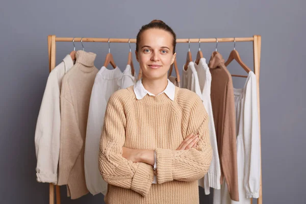 Indoor shot of calm confident woman with bun hairstyle wearing beige jumper standing against gray wall with clothes on hangers on shelf, keeps hand folded, looking at camera.