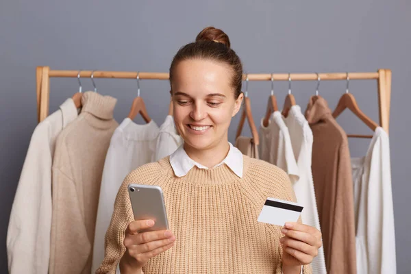 Beautiful happy woman wearing beige sweater, holding with credit card and cell phone, entering data and paying for shopping online, standing against gray wall with clothes hang in wardrobe on rack.