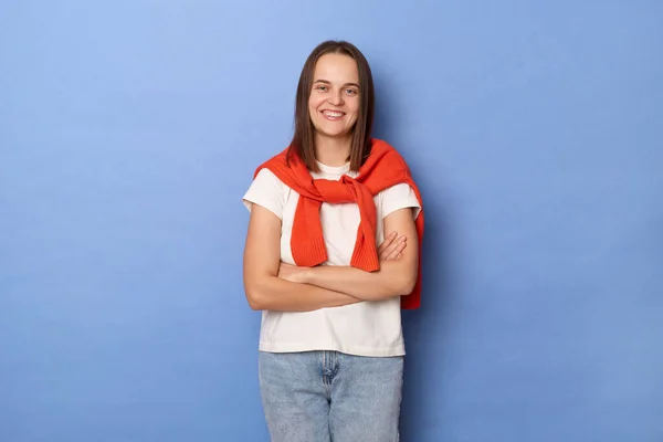 Woman wearing T-shirt and sweater tied over shoulders, smiling cheerfully, showing her white teeth to camera while feeling happy and carefree on her first day-off, posing isolated on blue background.