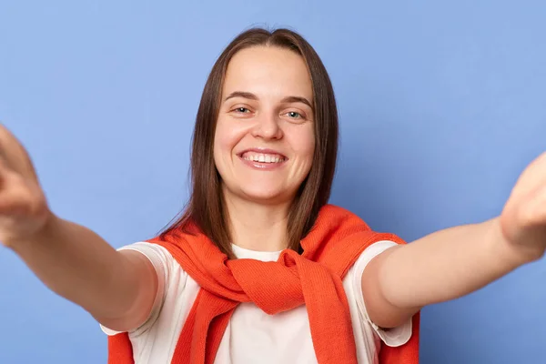 Portrait of smiling cheerful attractive dark haired woman wearing white T-shirt and orange sweater tied over shoulders, making selfie, point of view photo, posing isolated on blue background.