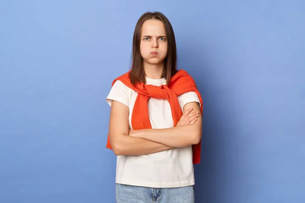 Indoor shot of sad upset Caucasian girl blows cheeks being offended with someone, female wearing white T-shirt and orange sweater tied over shoulders, standing against blue wall.