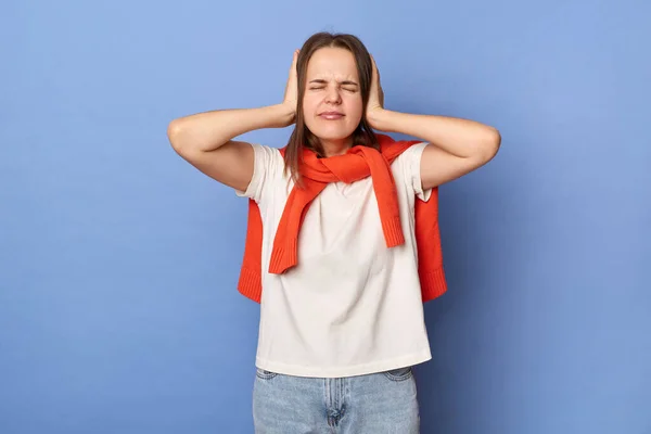 Portrait of despair dark haired woman wearing white T-shirt and orange sweater tied over shoulders, covering ears with hands, avoids noise, posing isolated on blue background.