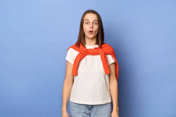 Indoor shot of surprised Caucasian girl wearing white T-shirt and orange sweater tied over shoulders, standing against blue wall, standing looking at camera with big eyes, sees something astonished.