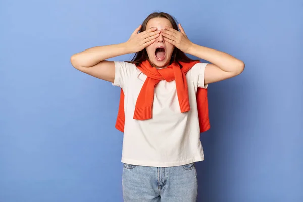 Image of shocked astonished surprised female with brown hair wearing white T-shirt and orange jumper tied over shoulders, standing isolated on blue background, dosen\'t want to see something shameful.