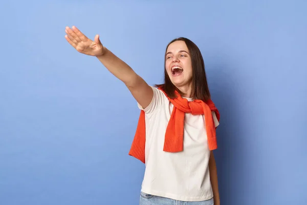 Photo of excited happy smile young adult woman dressed white T-shirt and orange jumper tied over shoulders standing against blue wall, standing with raised arms, screaming.