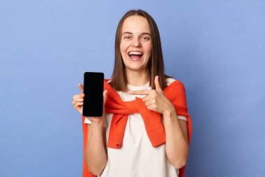 Portrait of happy positive woman in white T-shirt and orange sweater tied over shoulders, showing smart phone with empty display, pointing at her mobile phone, standing isolated on blue background.