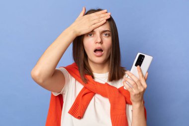 Photo of shocked young adult woman wearing white T-shirt and orange sweater tied over shoulders standing isolated on blue background, holding cell phone, touches her forehead, having problems.