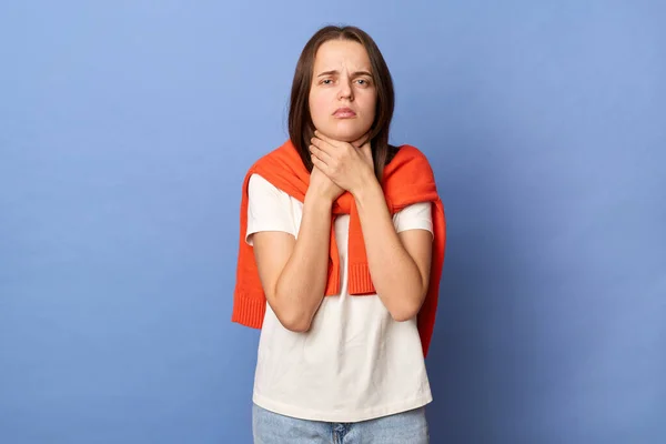 Photo of ill sick young adult woman wearing white T-shirt and orange sweater tied over shoulders, suffering sore throat, feels pain while swallowing, standing isolated on blue background.
