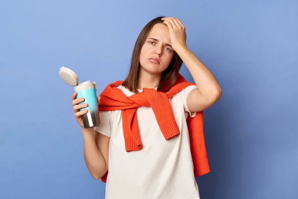 Image of sick tired Caucasian woman wearing white T-shirt and orange sweater tied over shoulders, holding thermos with coffee, suffering headache, standing isolated on blue background.