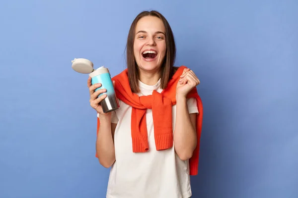 Portrait of extremely happy brown haired woman wearing white T-shirt and orange sweater tied over shoulders standing isolated on blue background, holding thermos with tea, clenched fist.