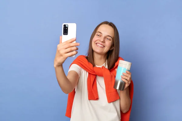 Image of smiling delighted Caucasian woman blogger wearing white T-shirt and orange sweater tied over shoulders standing isolated on blue background, taking selfie, making content for her blog.