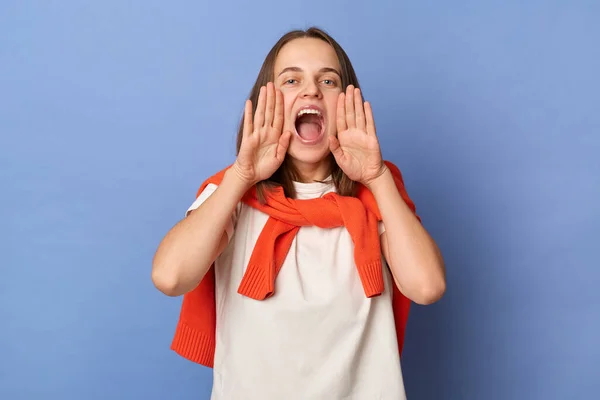 Indoor shot of beautiful attractive woman wearing white T-shirt and orange sweater tied over shoulders standing isolated on blue background, screaming loud with hands near mouth, making announcement.