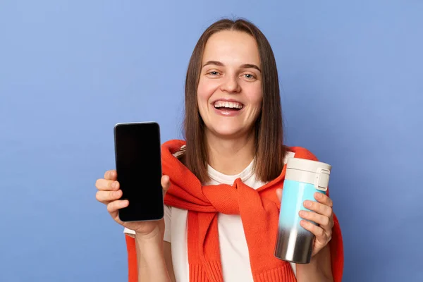 Portrait of cute smiling woman in white T-shirt and orange sweater tied over shoulders, showing empty display of her phone, holding thermos with coffee or tea, standing isolated on blue background.