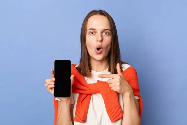 Indoor shot of shocked woman wearing white T-shirt and orange sweater tied over shoulders standing isolated on blue background, pointing at black blank screen of her phone, looking at camera.