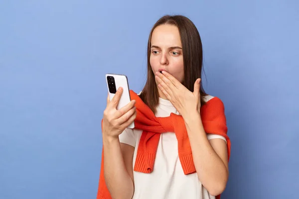 Shocked surprised amazed Caucasian woman wearing white T-shirt and orange sweater tied over shoulders, reading message on her phone, covering mouth with palm, standing isolated on blue background.