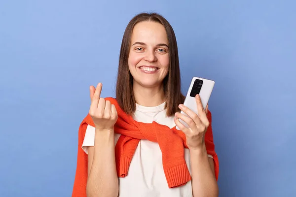 Image of charming smiling Caucasian woman wearing white T-shirt and orange sweater tied over shoulders standing isolated on blue background, using mobile phone, showing love gesture with fingers.