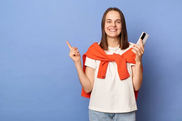 Smiling optimistic brown haired woman wearing white T-shirt and orange sweater tied over shoulders, holding mobile phone, pointing away ant copy space, standing isolated on blue background.