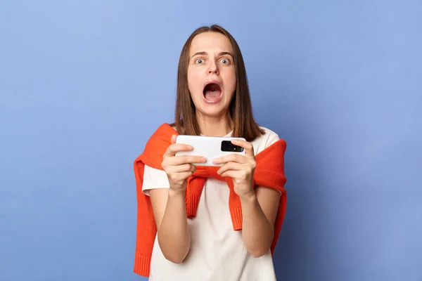 Portrait of shocked brown haired woman gamer wearing white T-shirt and orange sweater tied over shoulders standing isolated on blue background, loosing the level, screaming loud.
